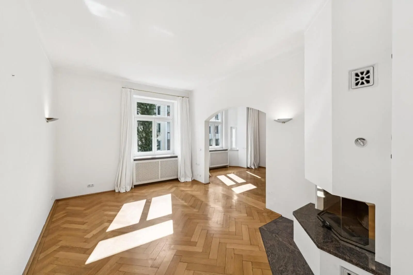 A bright, empty room with parquet flooring, white walls, large windows with white curtains, and a corner fireplace beside an arched doorway.