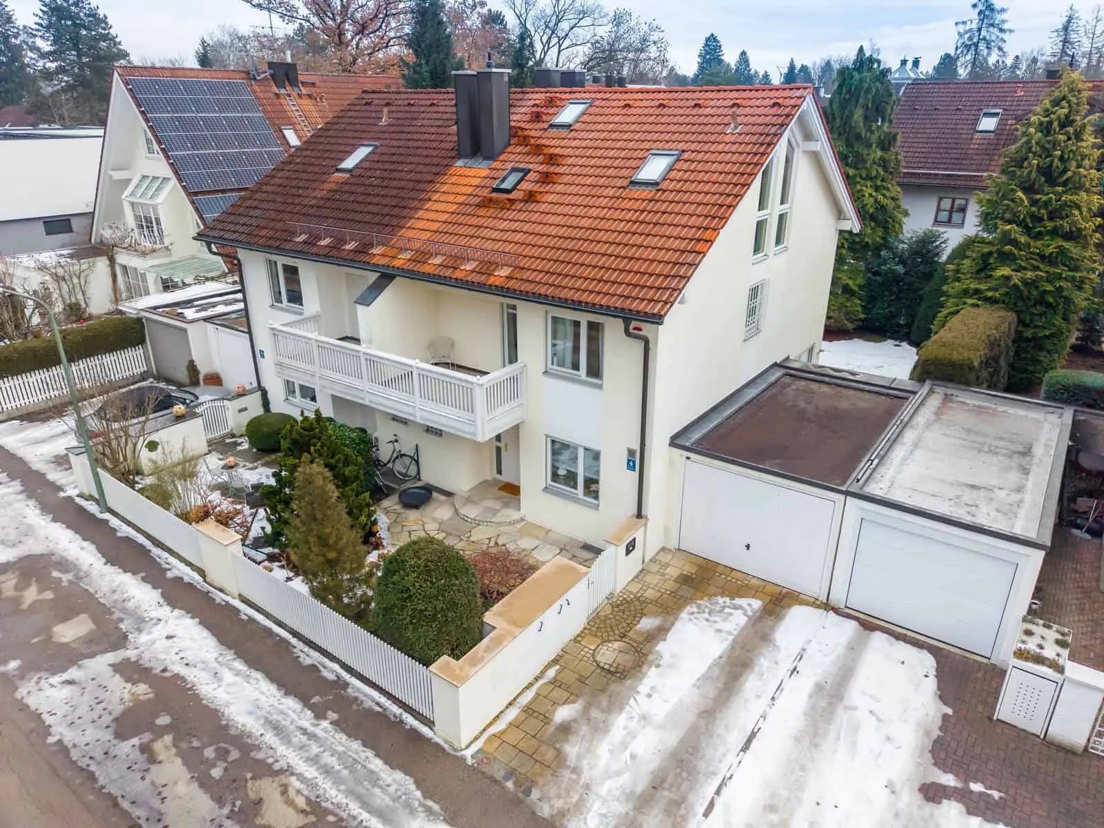 A two-story house with a red tile roof, white exterior, attached garage, small front garden, and patches of snow on the ground.