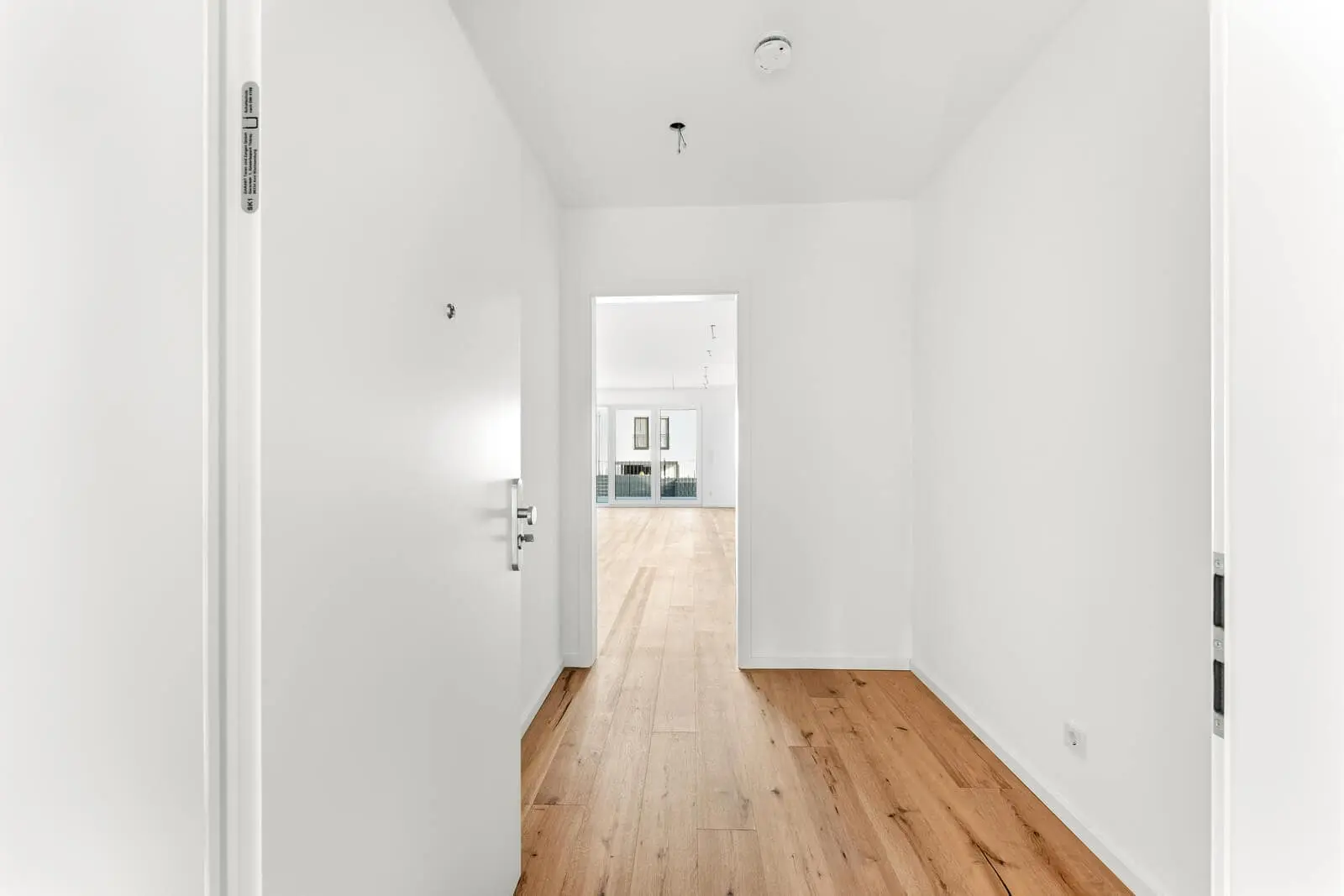 A bright, empty hallway with white walls and light wood flooring leads to a spacious room with large windows in the background.