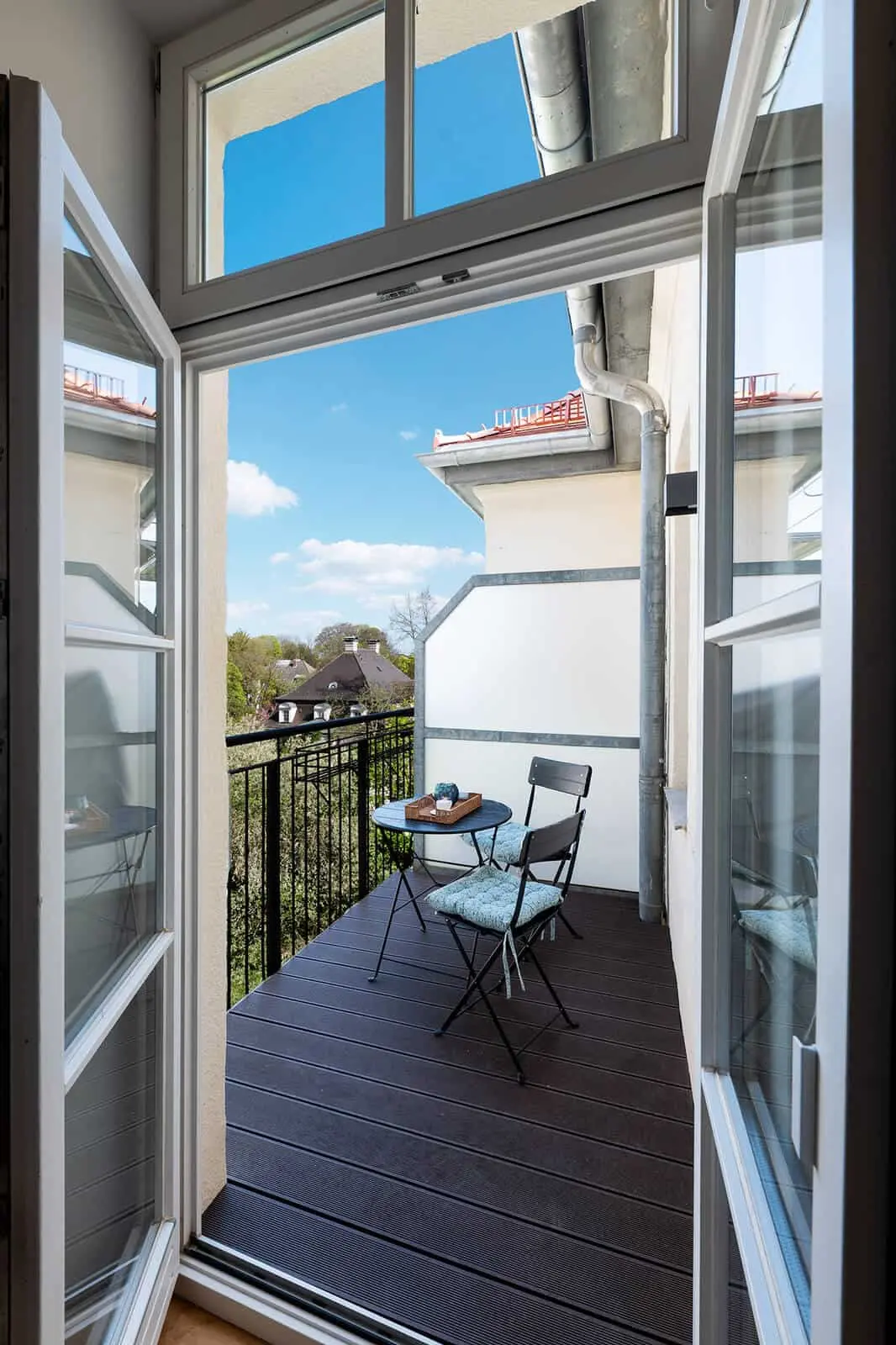 A small balcony with two black chairs and a round table set for coffee, viewed through open glass doors, with a scenic view of rooftops and trees under a blue sky.