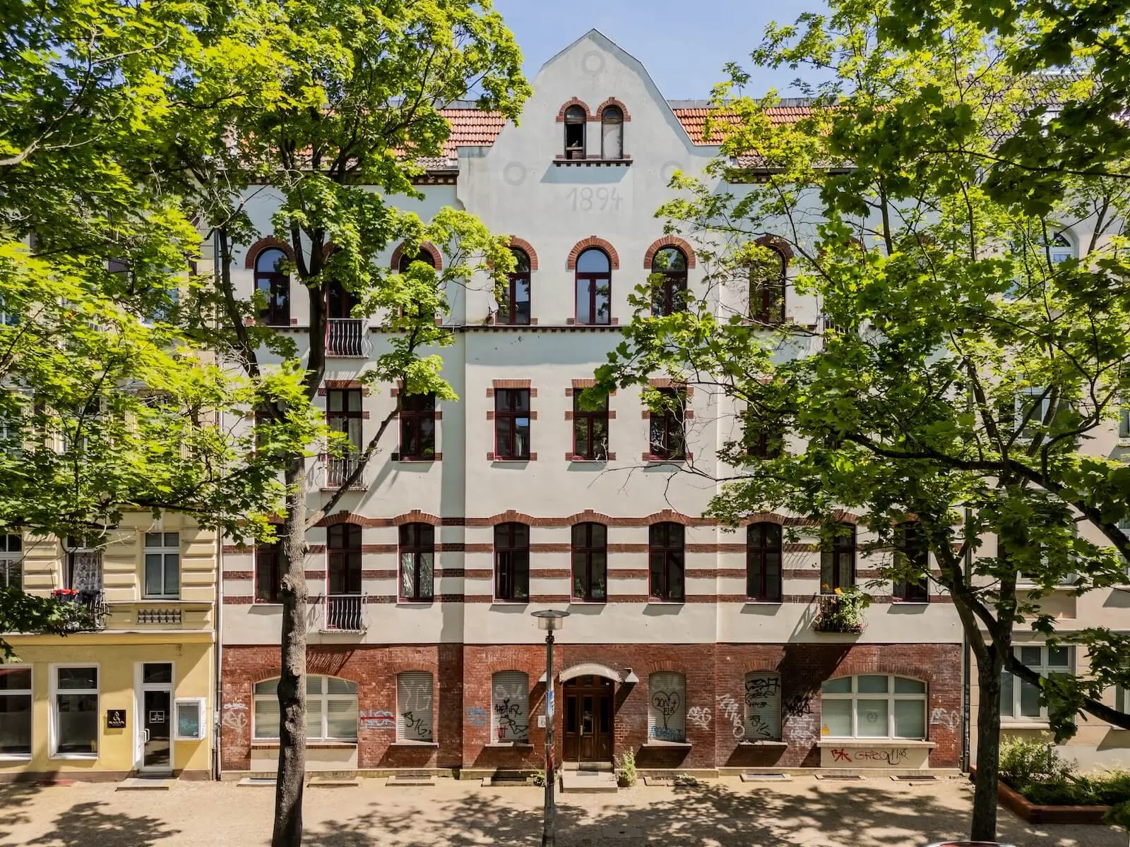 A four-story building with a painted year "1909" at the top, arched windows, and graffiti on the lower level, surrounded by leafy trees.