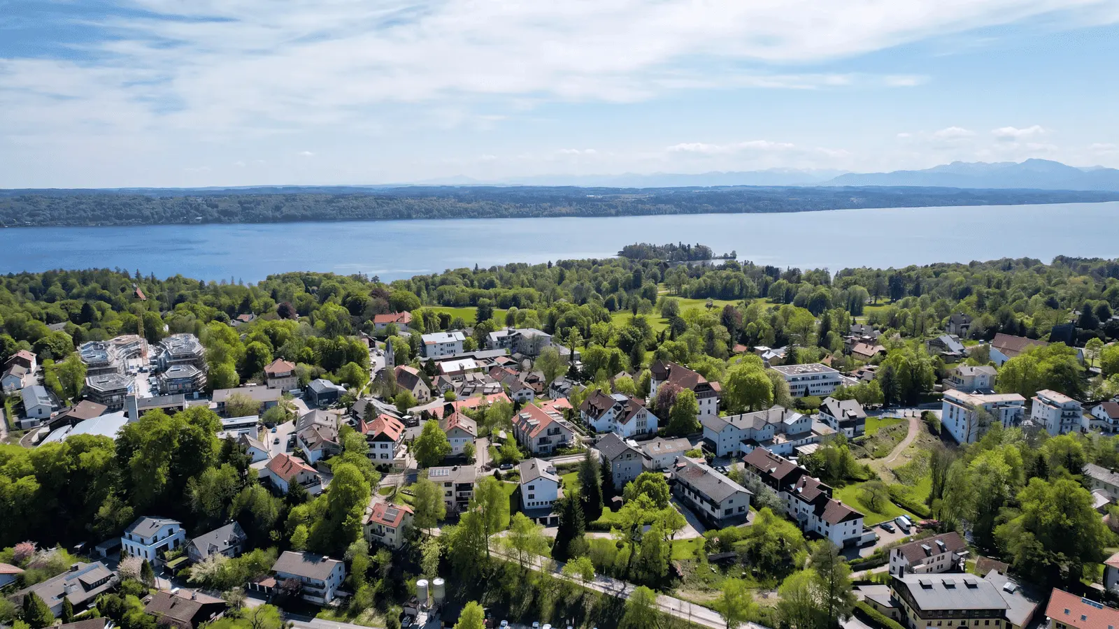 Aerial view of a small town with houses and trees near a large lake, with distant hills and a partly cloudy sky in the background.