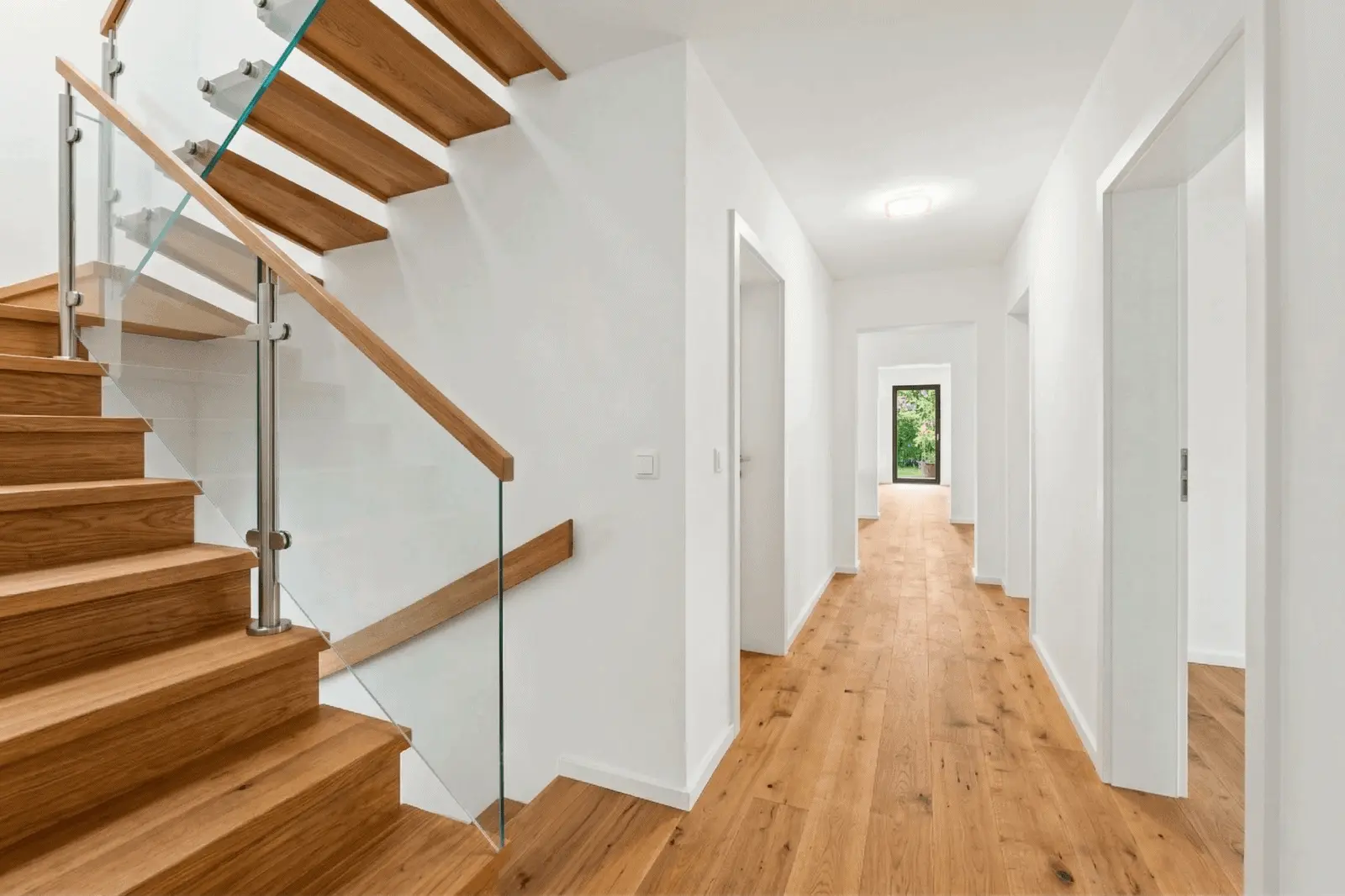 Modern hallway with wooden floors, white walls, and a staircase with wood steps and glass railing. Several doorways lead to other rooms; a window at the end shows greenery outside.