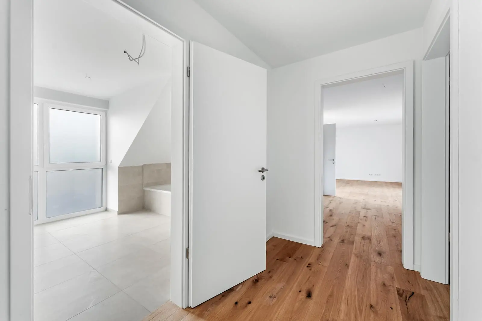 Empty hallway with wooden floors, white walls, and open doors leading to a tiled bathroom and a bright, unfurnished room with large windows.