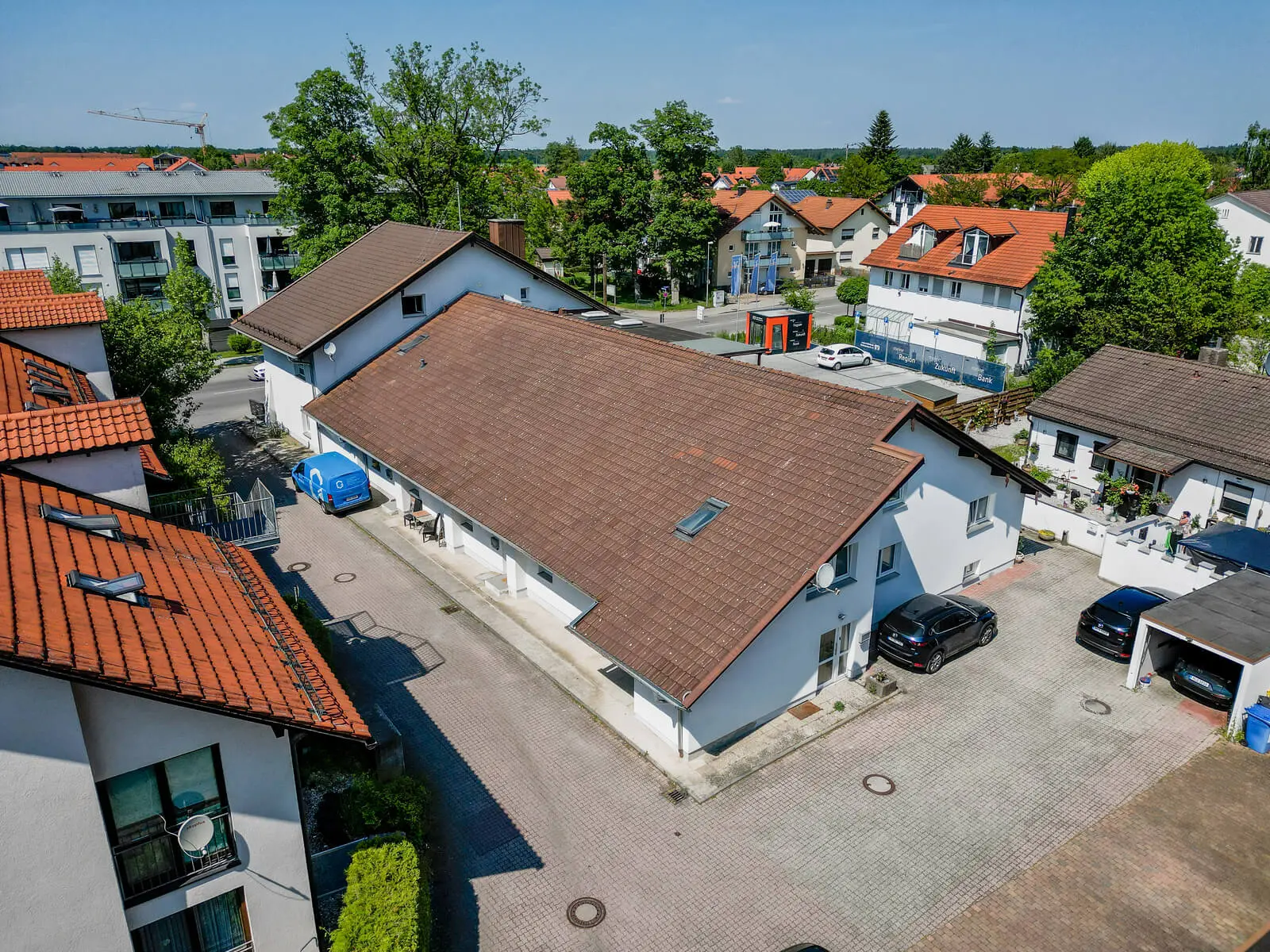 Aerial view of a residential area featuring several houses with red-tiled roofs and parked cars, surrounded by greenery and other buildings.