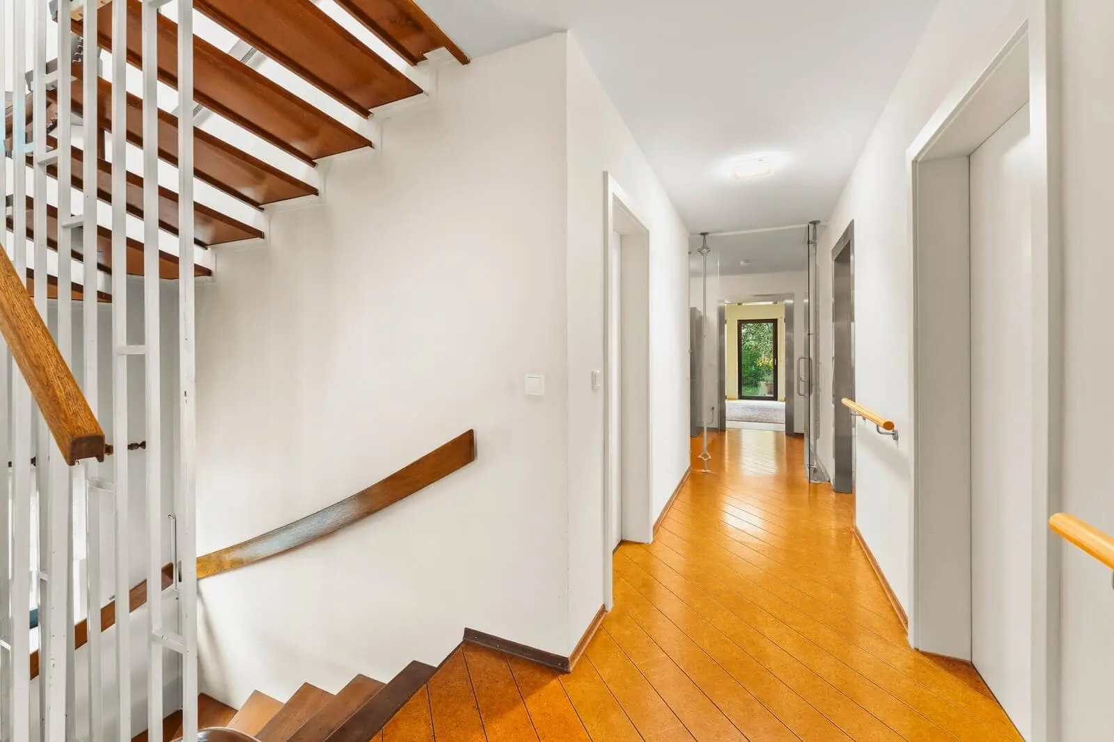 A bright hallway with wooden floors, white walls, multiple doorways, and a wooden staircase with metal railings leading to an upper level.