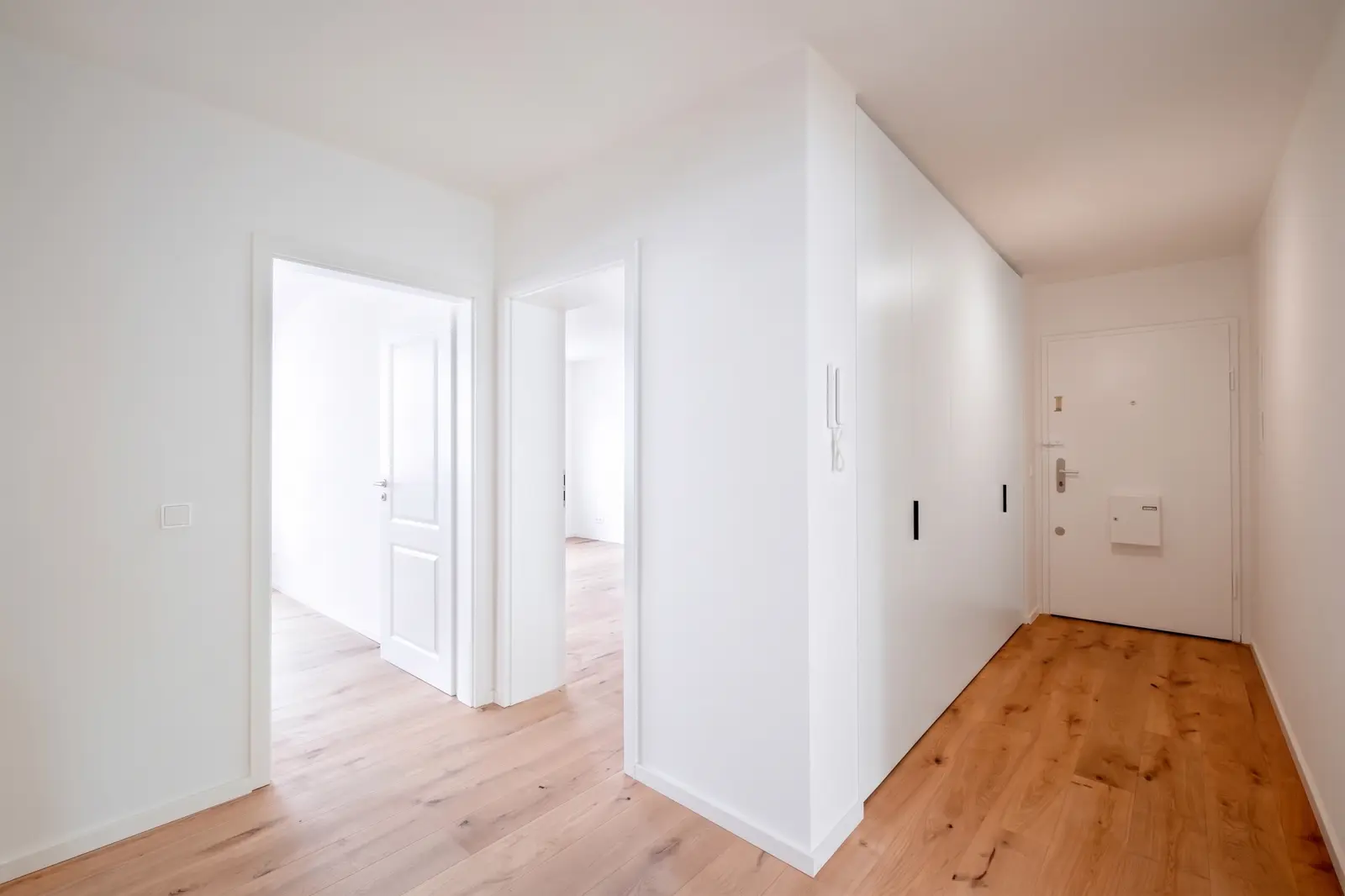 Heller Flur in einer modernen Wohnung mit weißen Wänden, offenen Türen zu den angrenzenden Räumen und durchgehend hellem Holzfußboden.