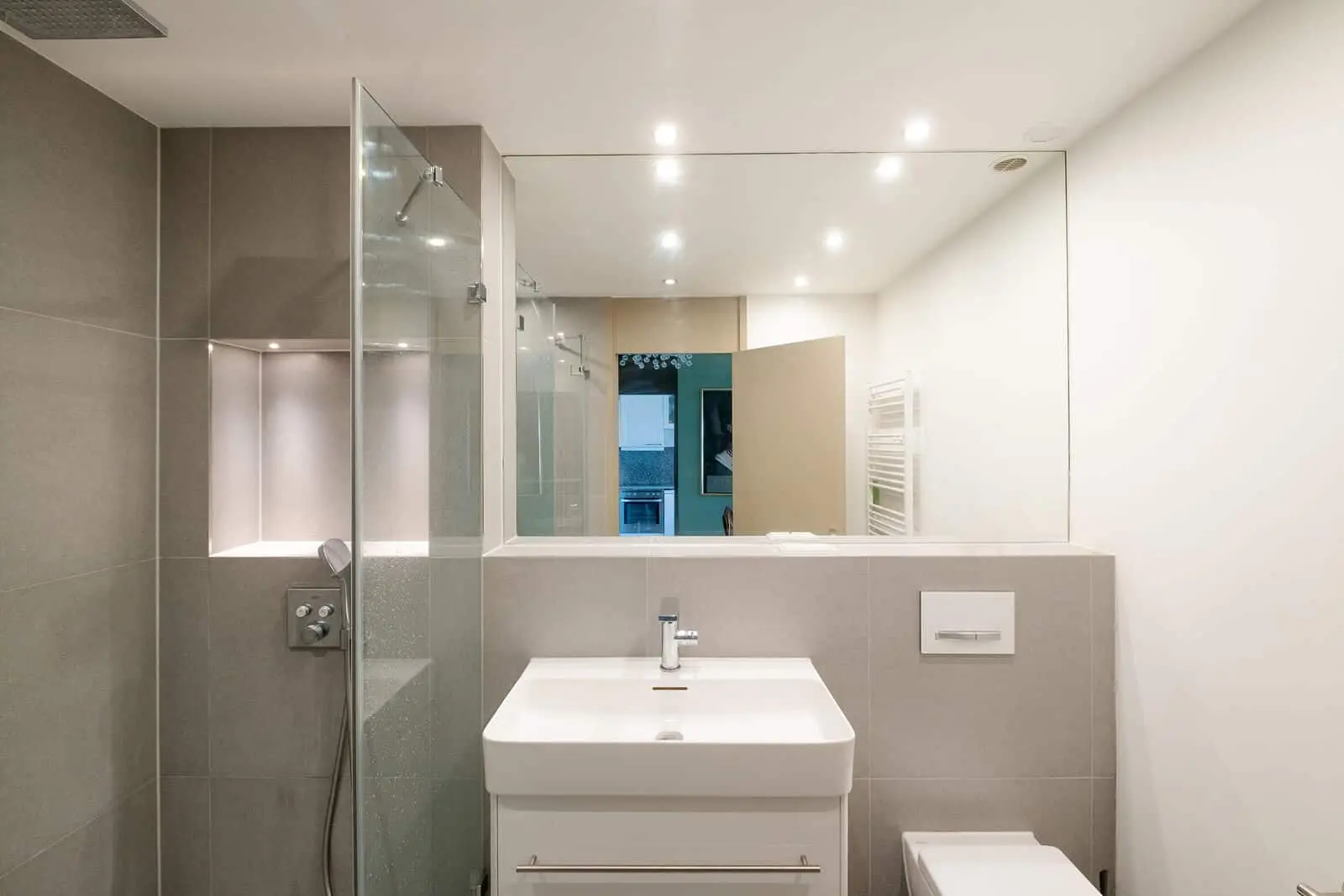 Modern bathroom with gray tiled shower, glass partition, white sink with cabinet, wall-mounted faucet, large mirror, and recessed lighting.