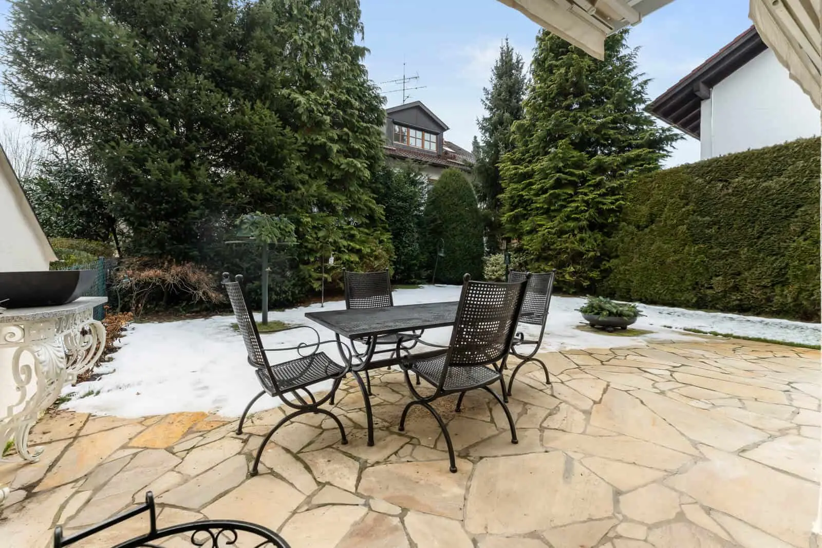 A metal patio table with four chairs sits on a stone terrace in a backyard bordered by evergreen trees, with patches of snow on the ground.
