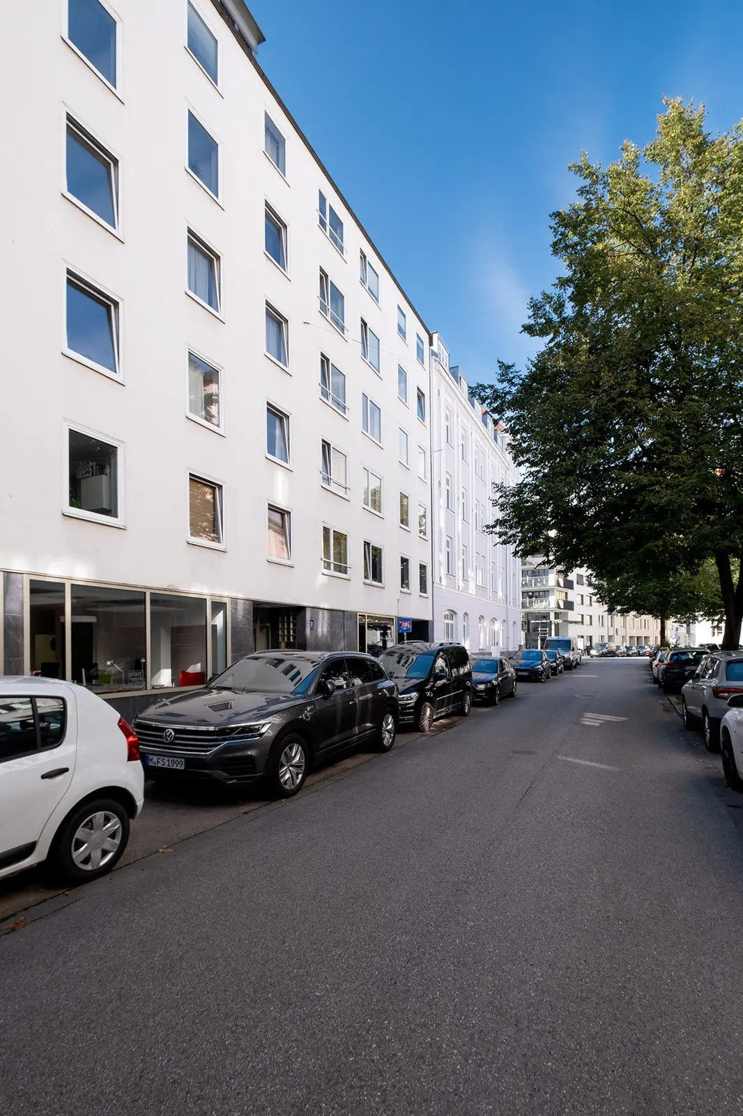 Eine Reihe geparkter Autos säumt eine schmale Straße neben weißen Mehrfamilienhäusern mit vielen Fenstern und einem Baum auf der rechten Seite.