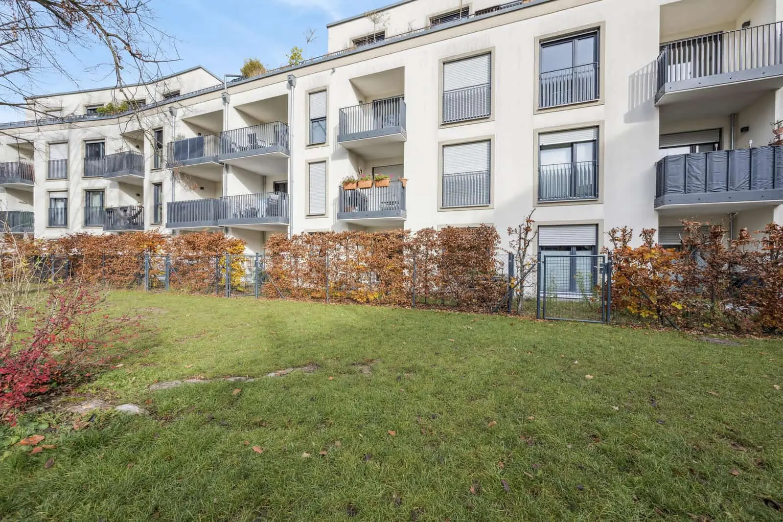 Modernes Mehrfamilienhaus mit Balkonen und großen Fenstern, mit Blick auf eine grüne Wiese, die von einer Hecke und einem Metallzaun begrenzt wird.