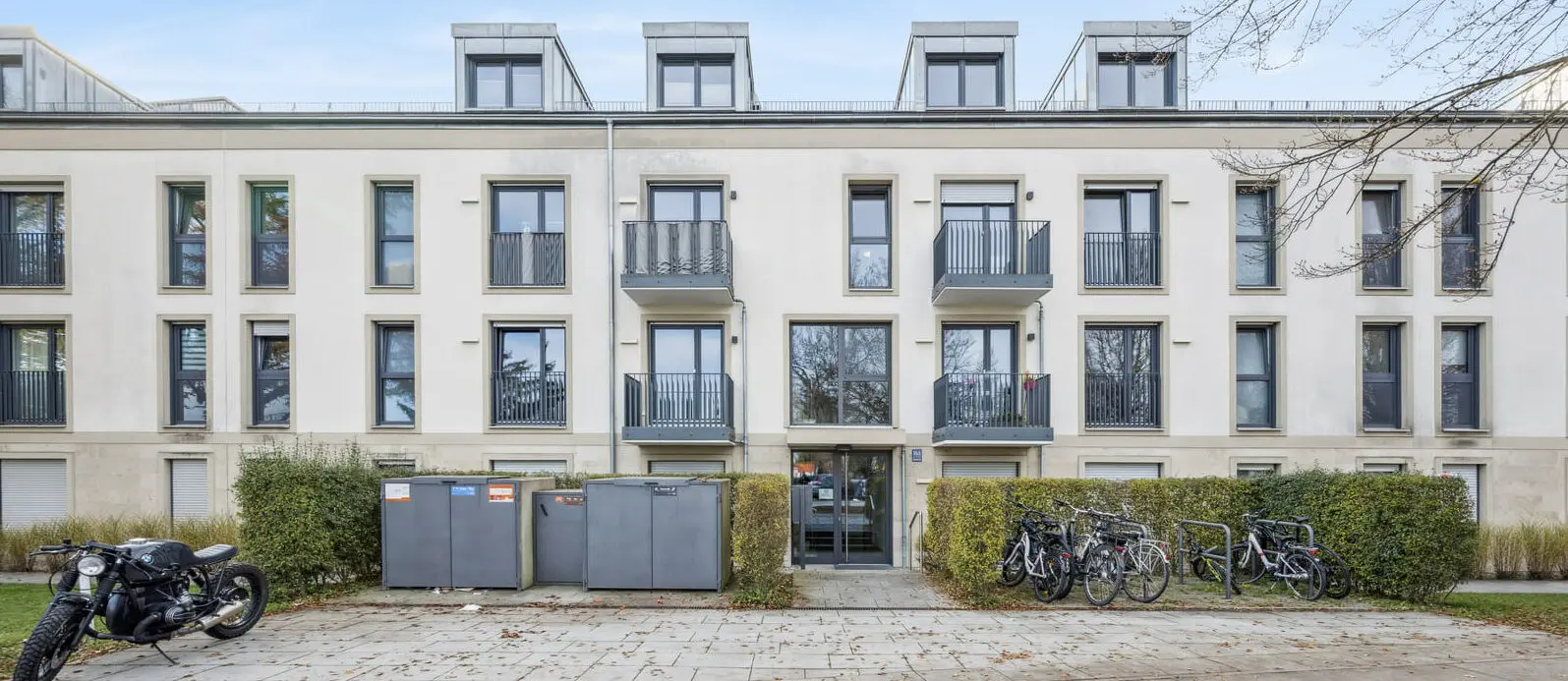 A modern three-story apartment building with balconies, bike racks, waste bins, and a motorcycle parked in front. Fallen leaves cover parts of the pavement and ground.