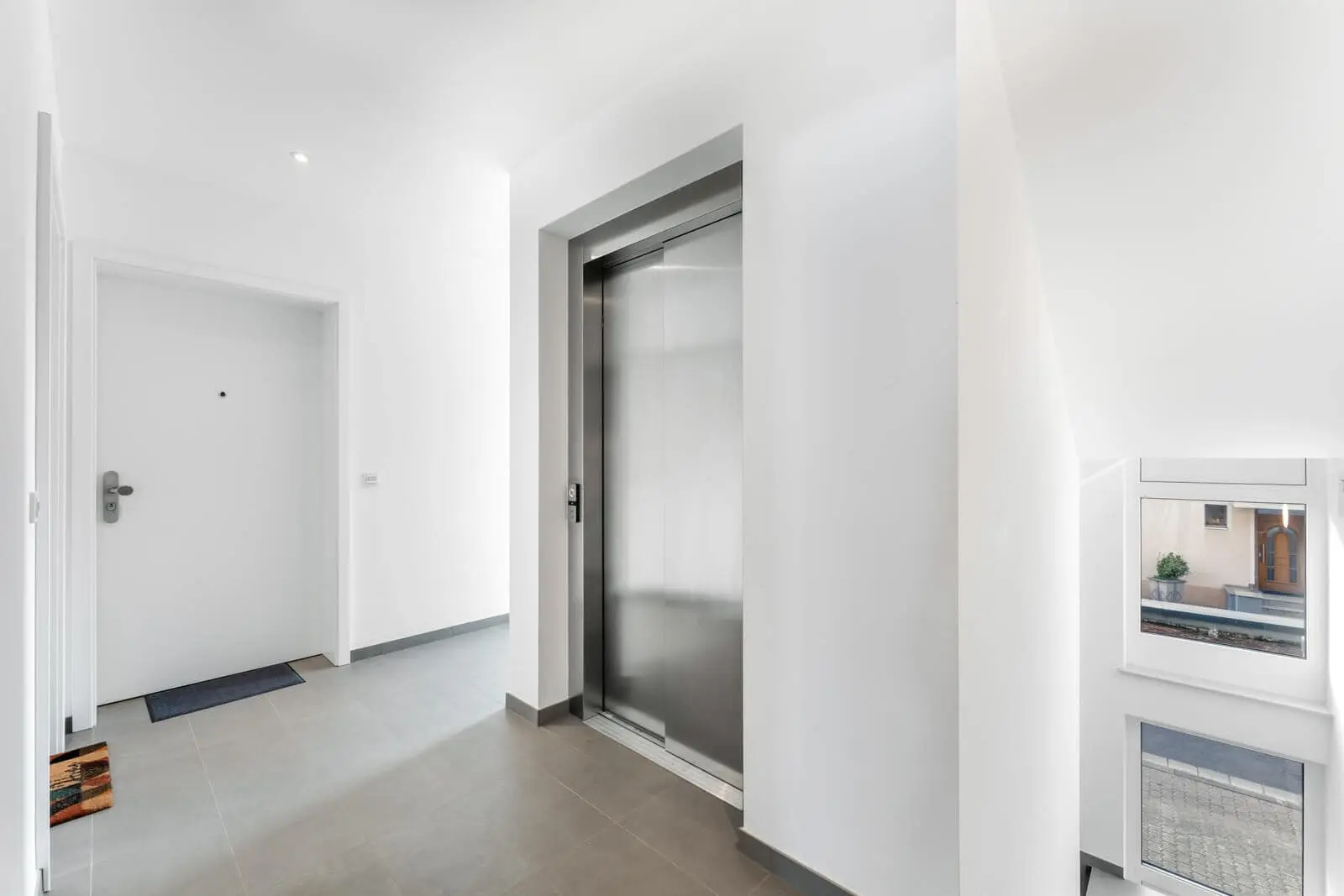 Modern apartment building hallway with a closed elevator door, a white entrance door, gray tile flooring, and a window near a stairwell.