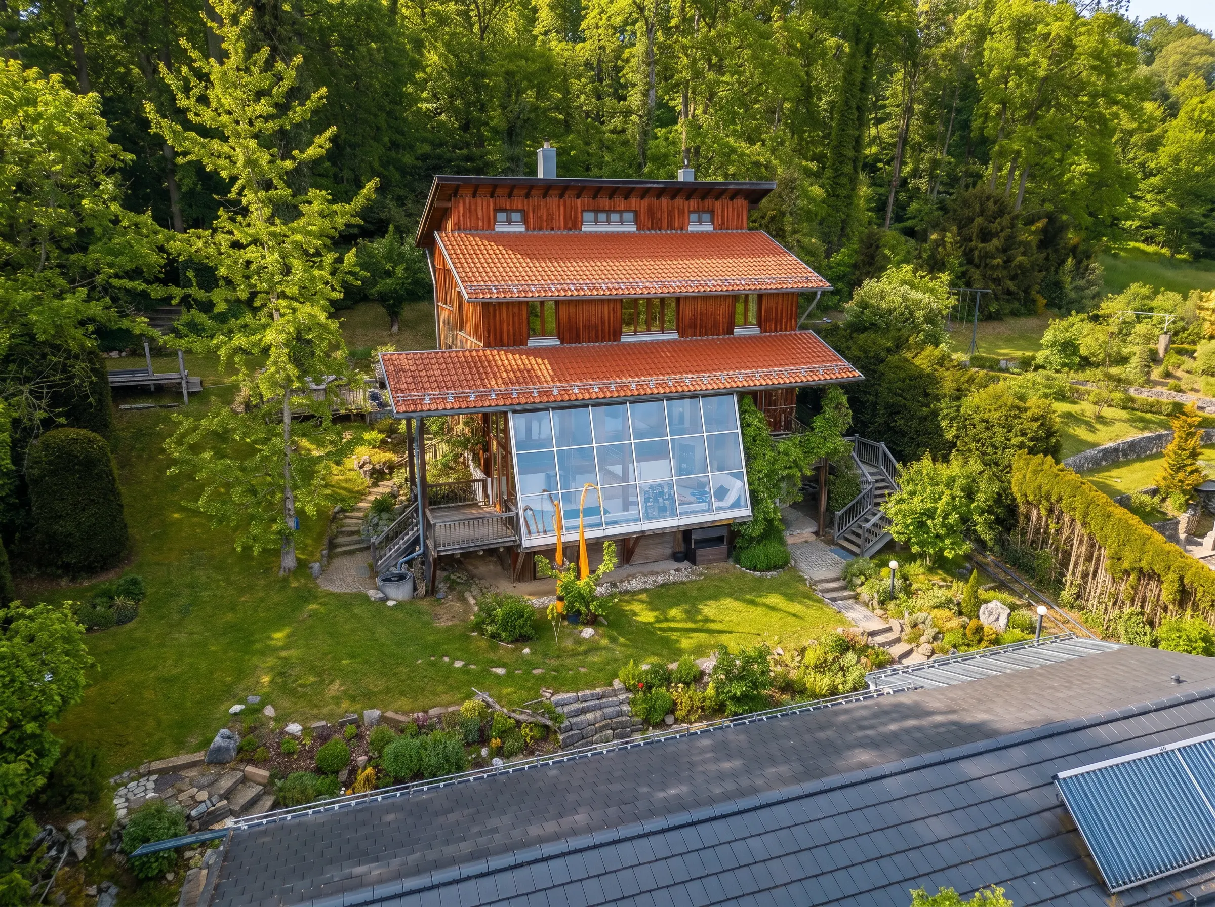A timber house with a red tiled roof and a large glazed terrace sits amid lush vegetation and well-kept gardens in a wooded area.