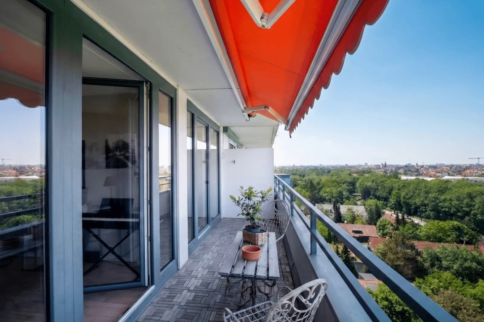 A modern apartment balcony with a red retractable awning, glass doors, outdoor furniture, potted plant, and a view of trees and city buildings in the distance under a clear sky.