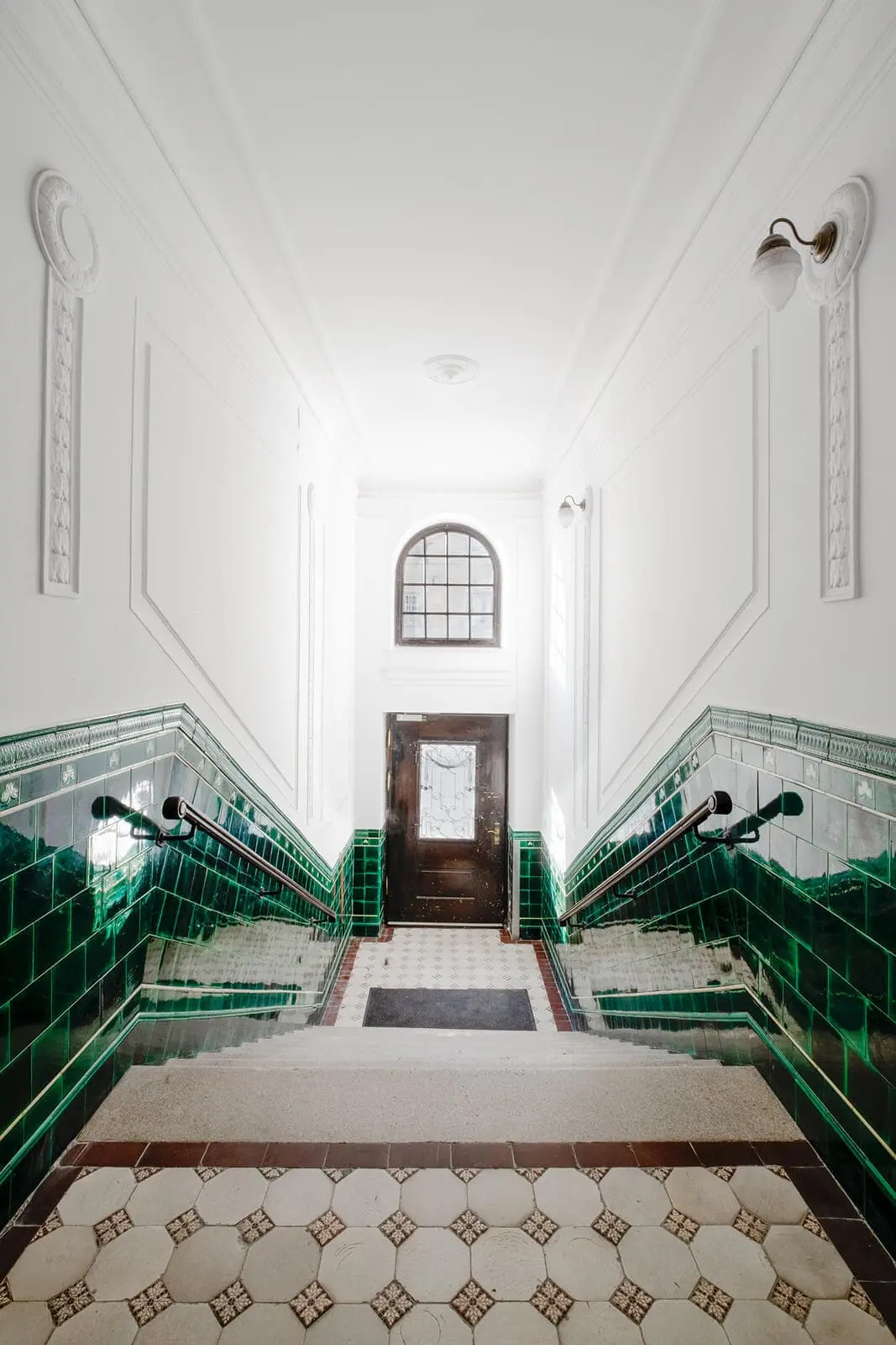 A stairwell with green tiled walls, white decorative molding, and a brown door with a window at the bottom, lit by natural light from a high arched window.