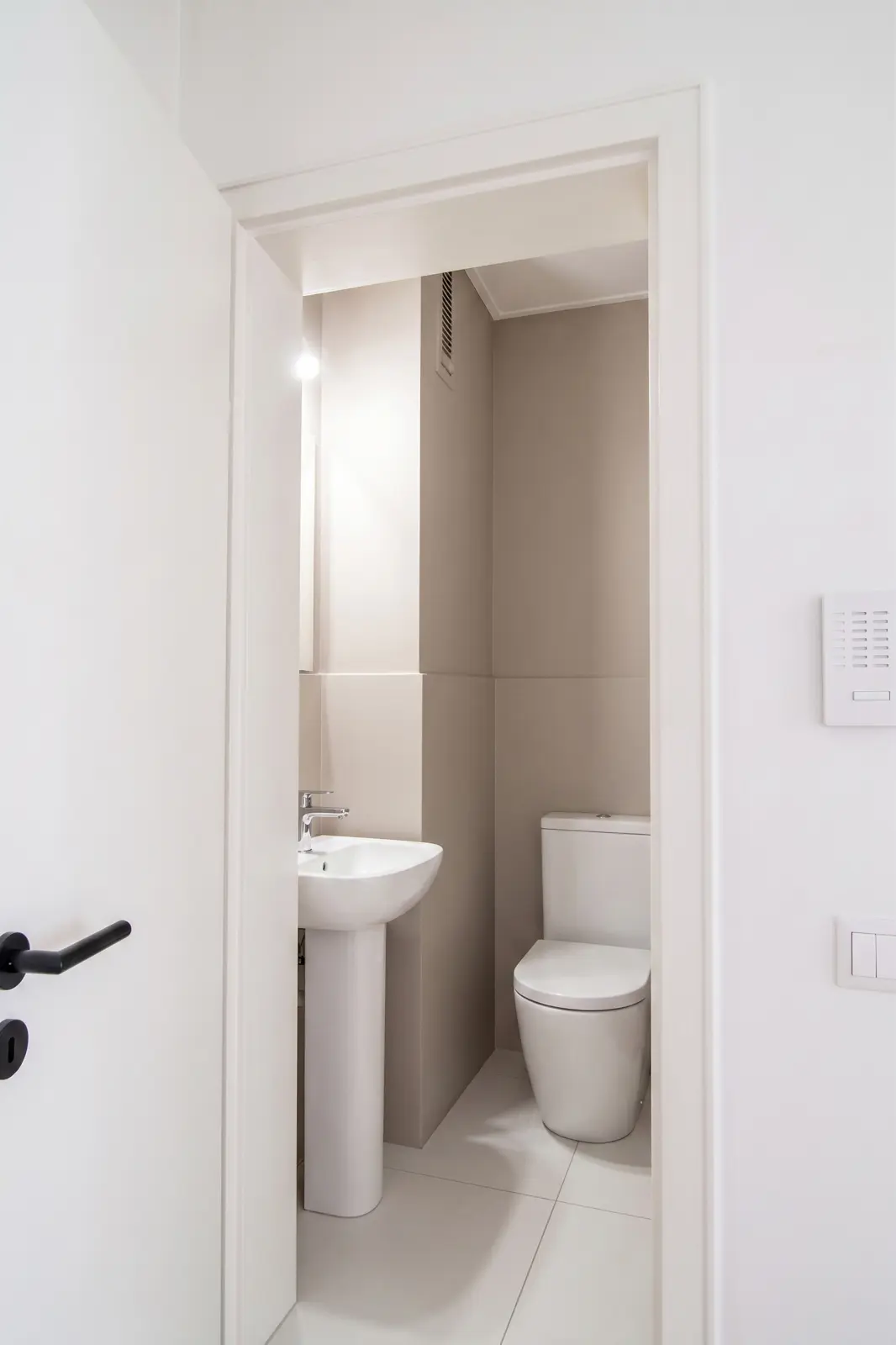 Small, modern bathroom with a white toilet and a pedestal washbasin, beige walls and a partly open white door.