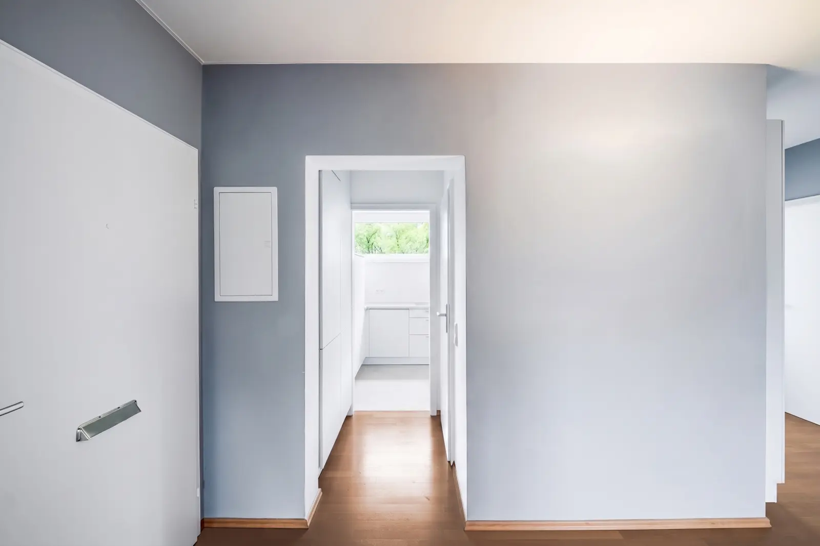 A hallway with light blue walls and wooden floors leads to a bright white kitchen whose window looks out onto the green landscape outside.