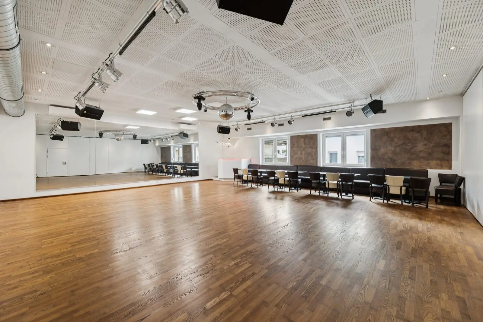 A spacious, empty dance studio with a wooden floor, large mirrors on one wall, ceiling lights, a disco ball and chairs lined up against the opposite wall.