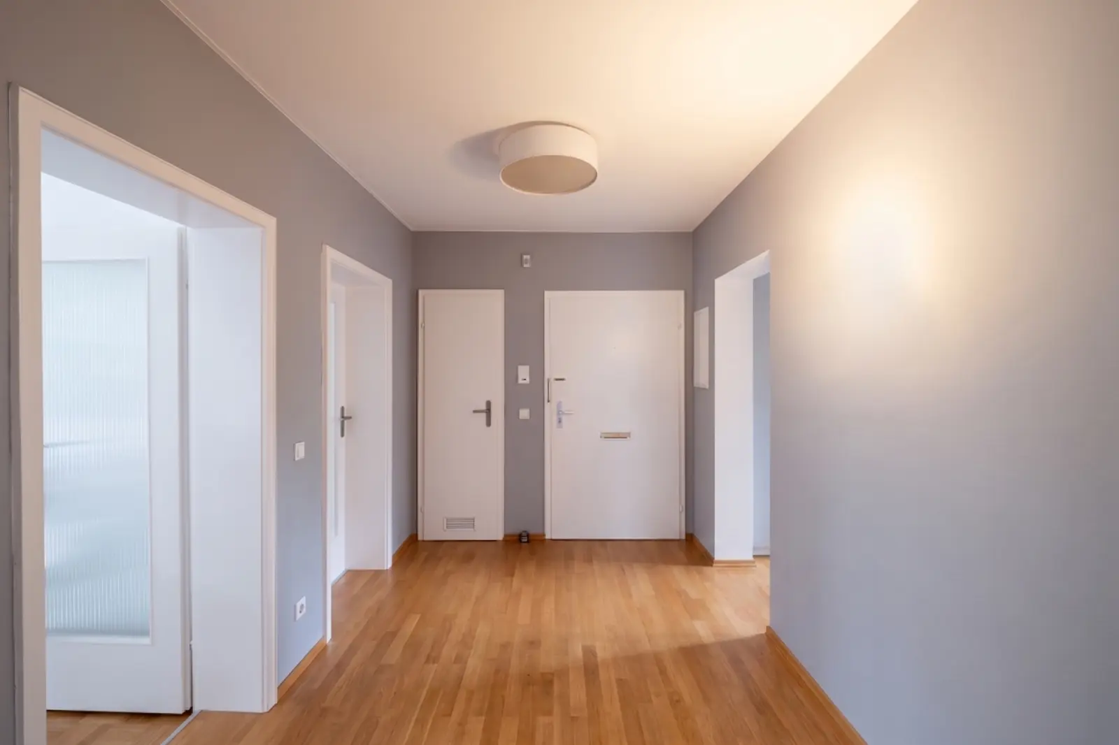 An empty hallway with light grey walls, a wooden floor, white doors and a round ceiling light. The space is well lit by daylight coming in from an adjoining room.