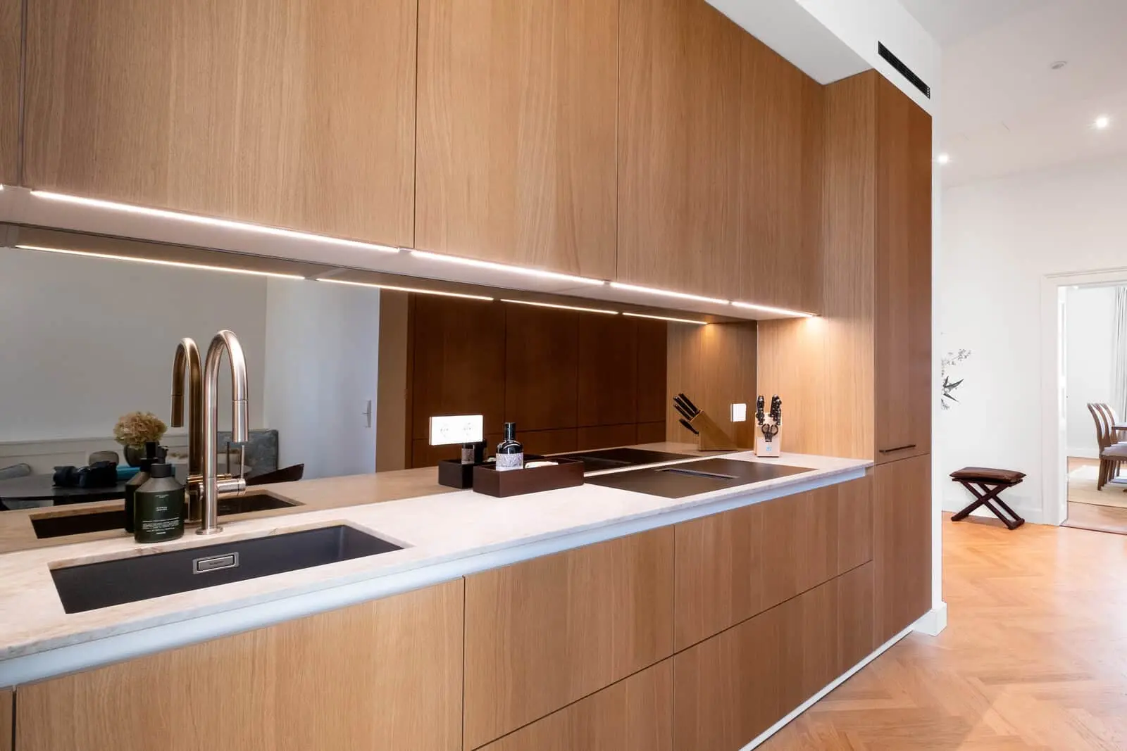 Modern kitchen with wooden cabinets, under-cabinet lighting, two sinks with chrome faucets, marble countertop, and minimalist decor. Dining area visible through open doorway.