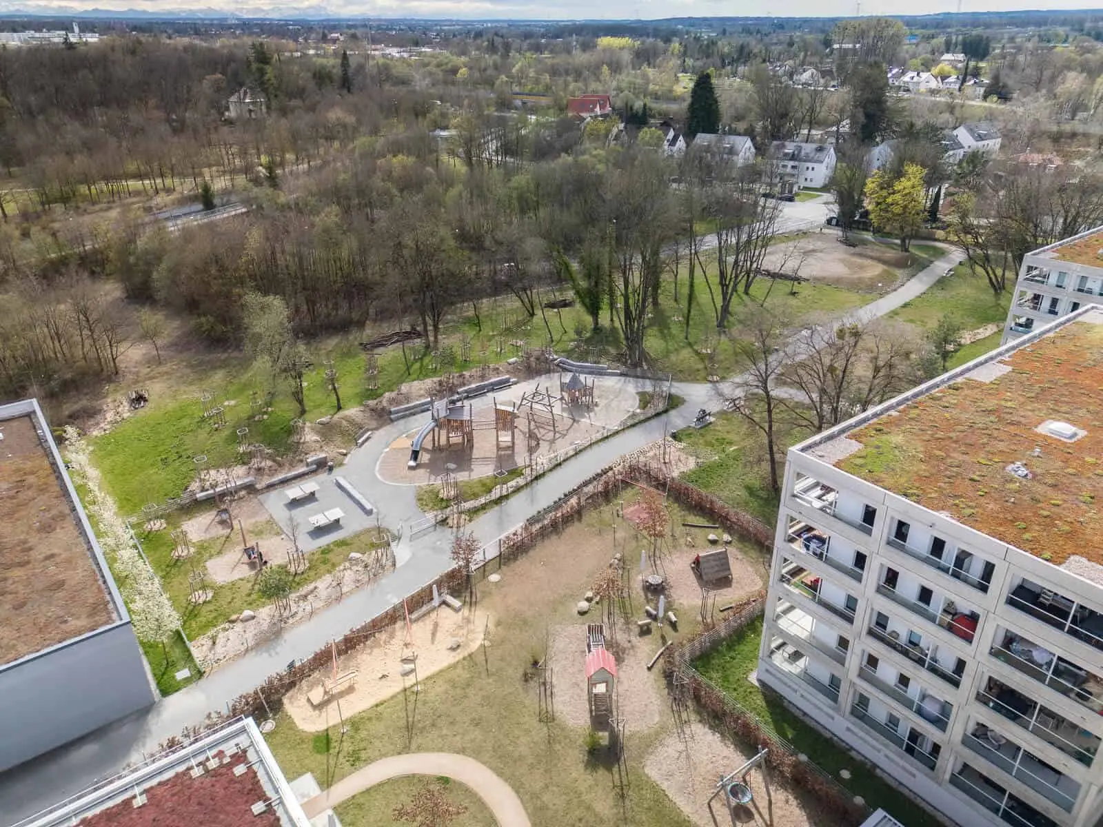 Aerial view of a residential area with apartment buildings, playgrounds, walking paths, and surrounding trees and grassy spaces.