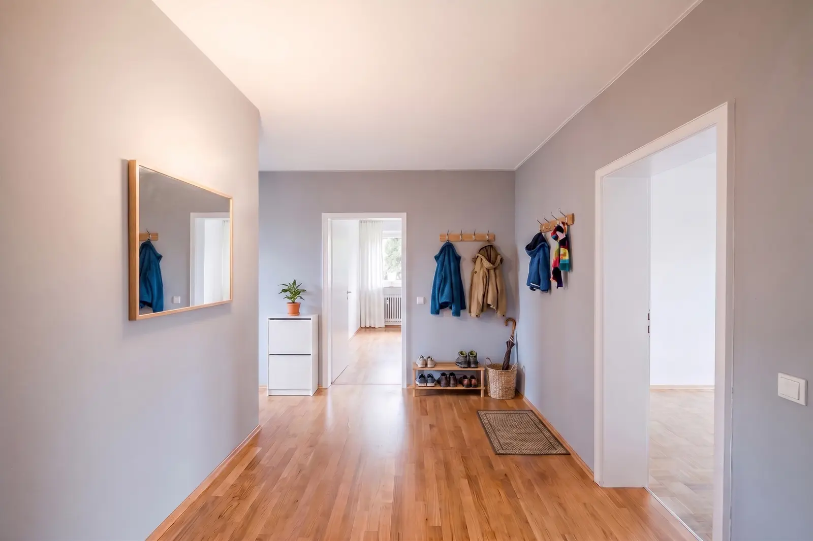 A tidy hallway with a wooden floor, coat rails with jackets, a shoe rack, a mirror on the wall, a small cabinet with a plant and doors leading to other rooms.