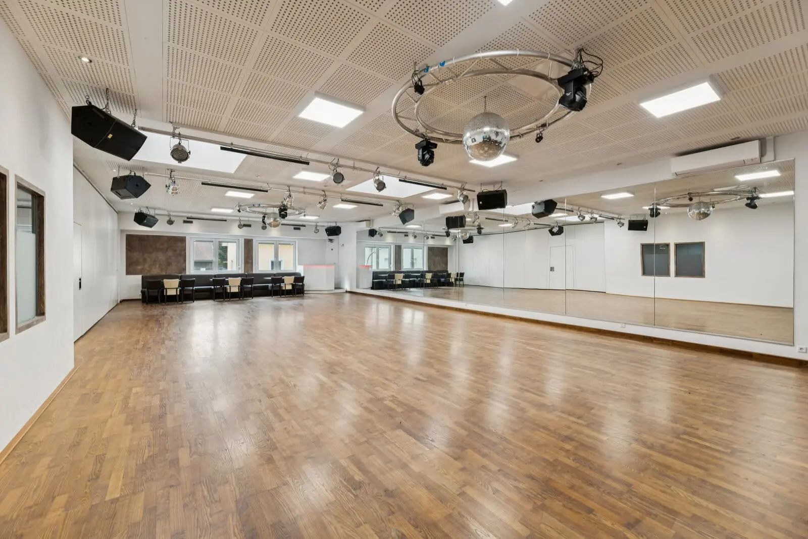 Spacious empty dance studio with a wooden floor, large mirrors on one wall, windows, ceiling lights, speakers and a disco ball.