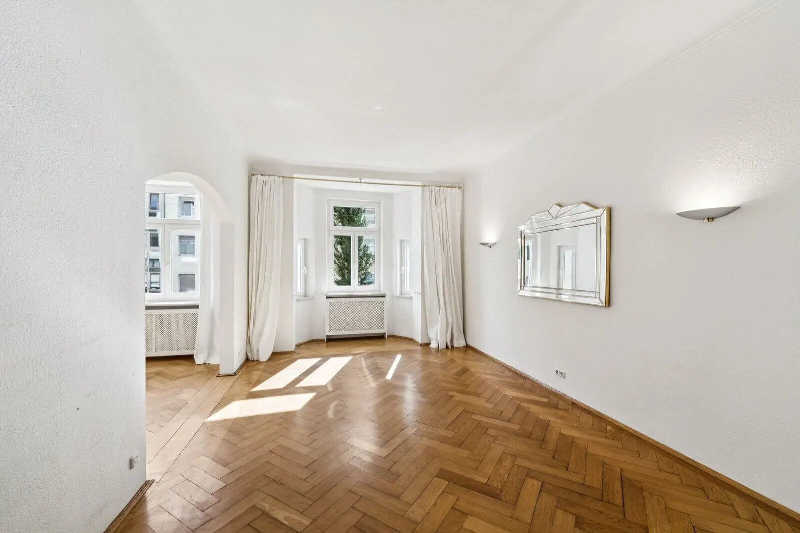 Bright, empty room with parquet flooring, white walls, a large mirror, wall sconces, and three windows with white curtains letting in natural light.