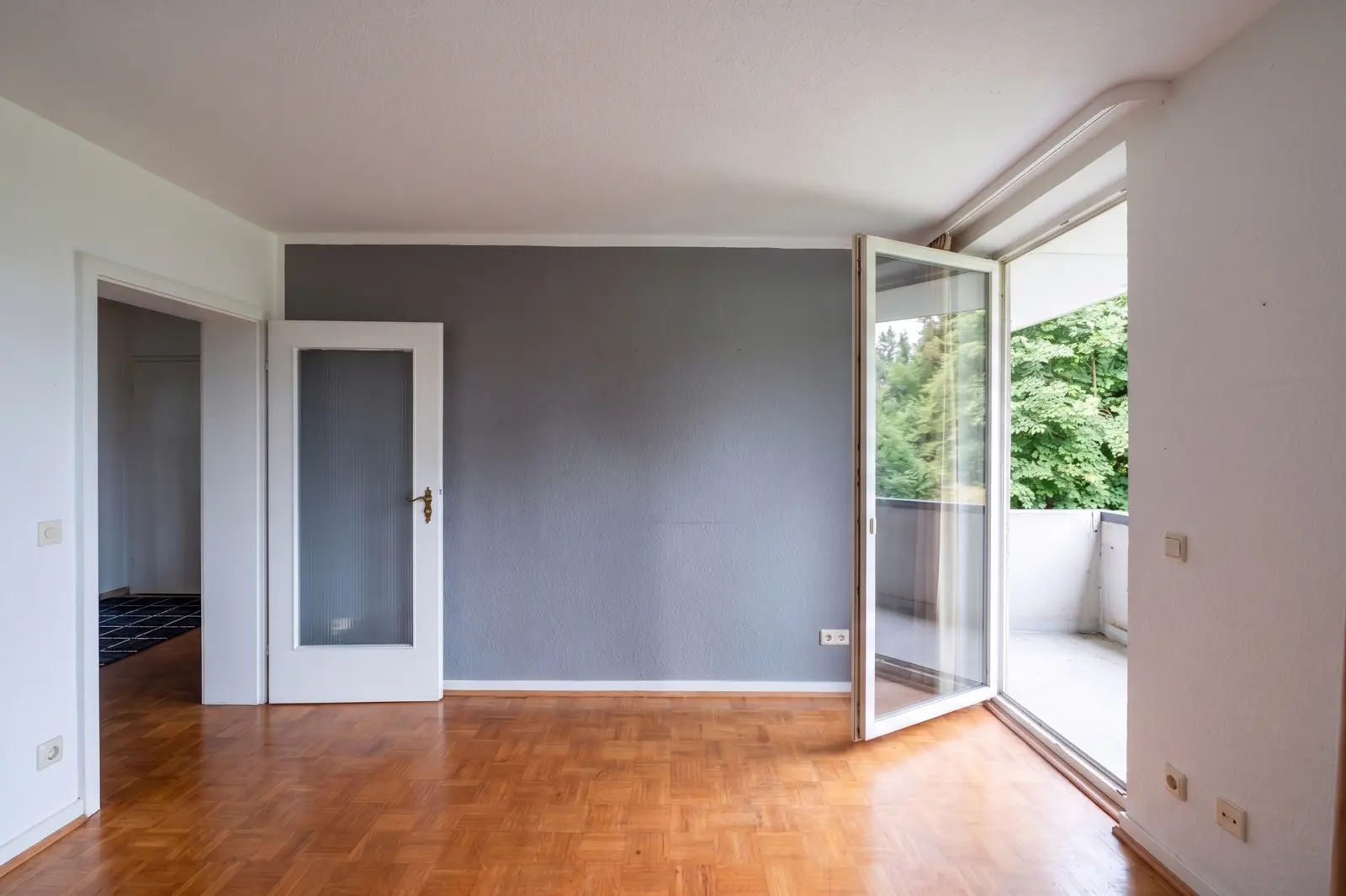 Empty room with parquet flooring, a light grey accent wall, an open door to the hallway and an open glass door leading to a balcony with a view of greenery.