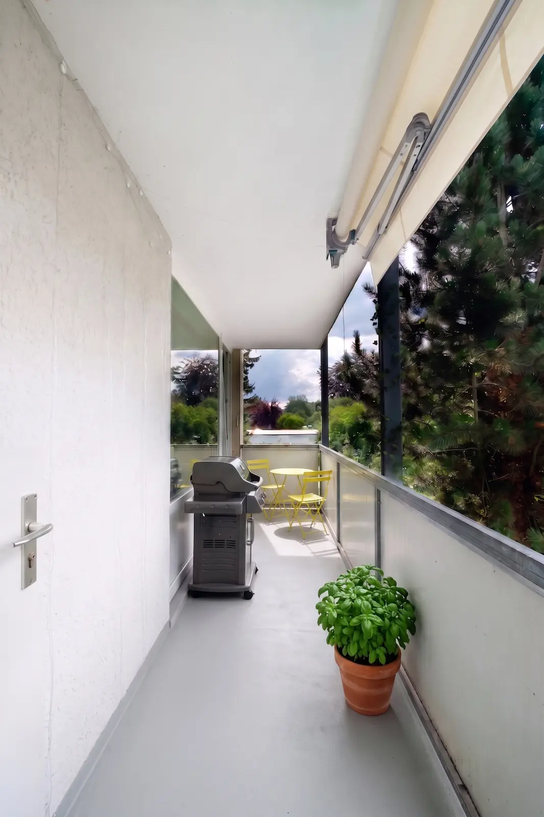 A narrow balcony with a barbecue, a potted basil plant and a small yellow table with two chairs, looking out through a glass balustrade onto green surroundings.