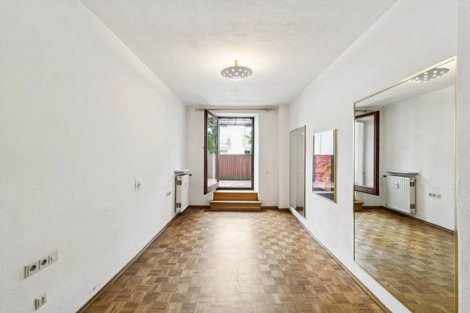 Empty room with white walls, parquet flooring, large wall mirror, radiator, and an open window leading to a small outdoor balcony area.