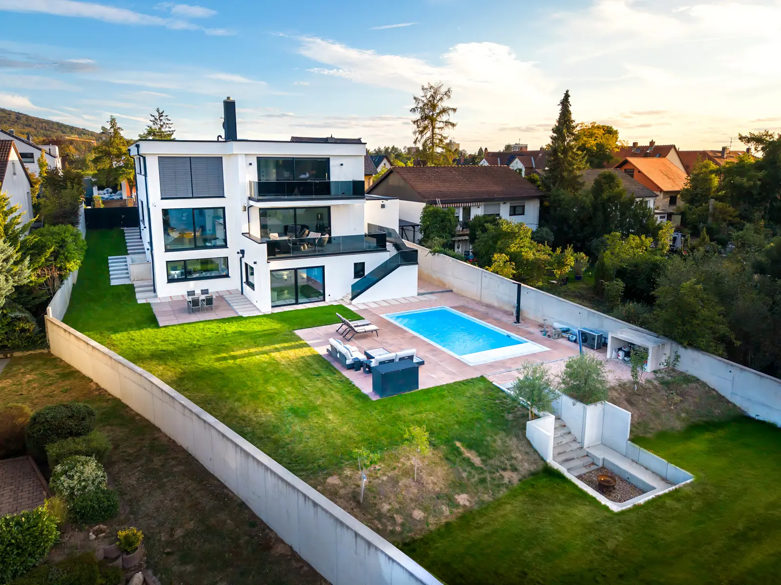 Aerial view of a modern white multi-story house with a swimming pool, patio furniture, and a lawn enclosed by a perimeter wall.