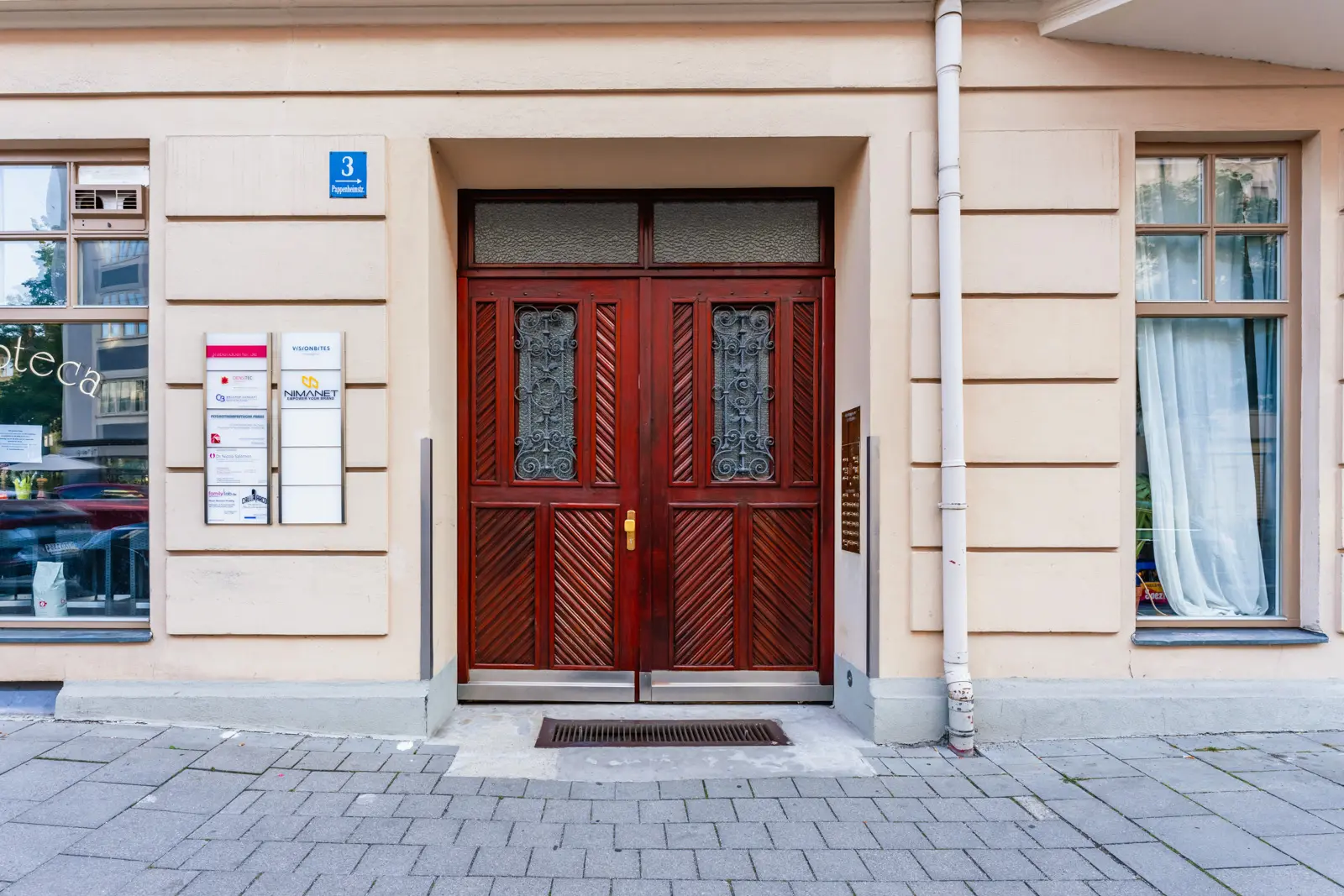 Double wooden doors with decorative iron panels in a beige building, flanked by windows; address plaque and business signs on the left.