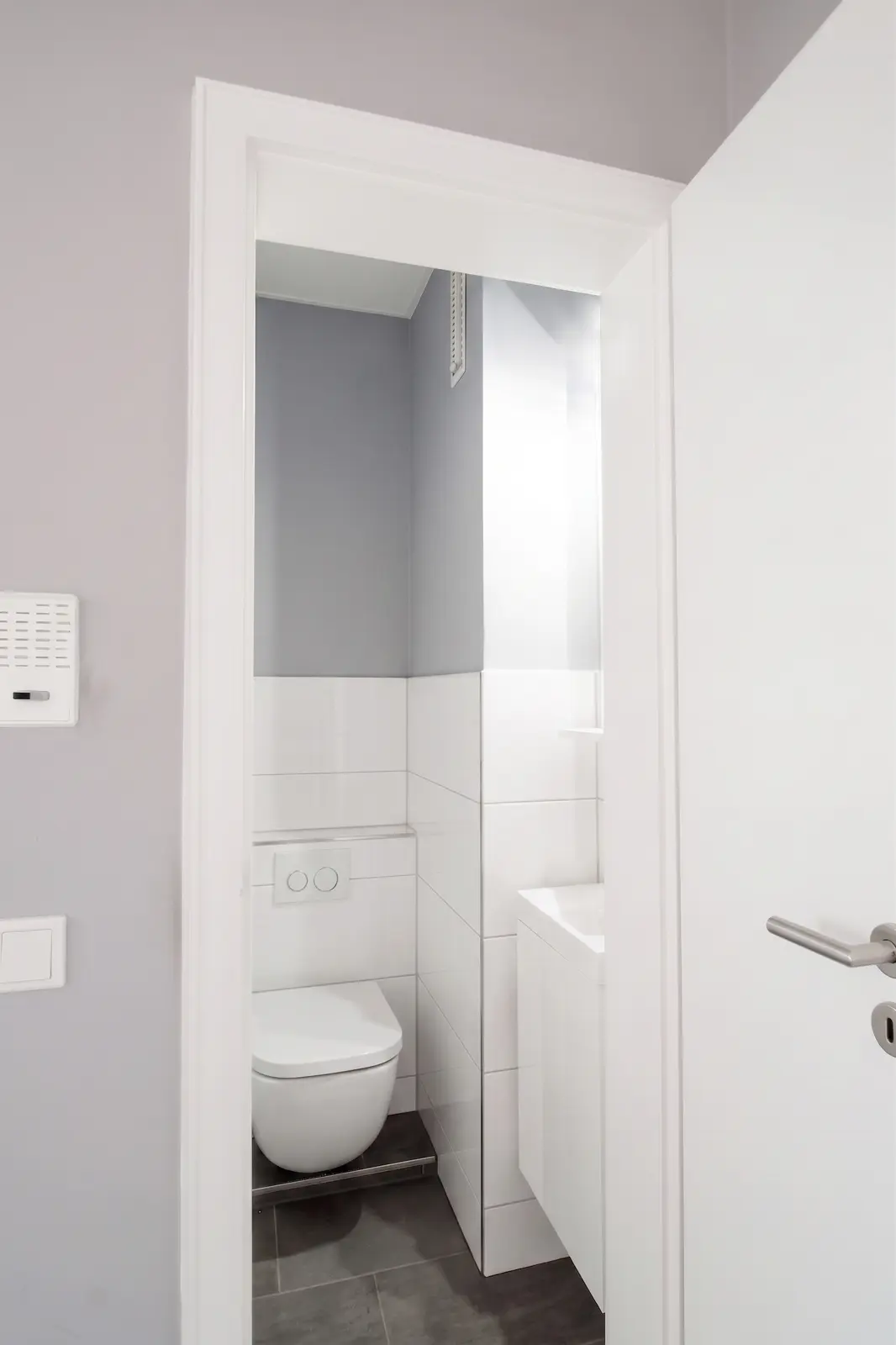 Small, modern bathroom with white tiles, a wall-hung toilet, a compact washbasin and a partly open door. The room has light grey and white walls and dark floor tiles.