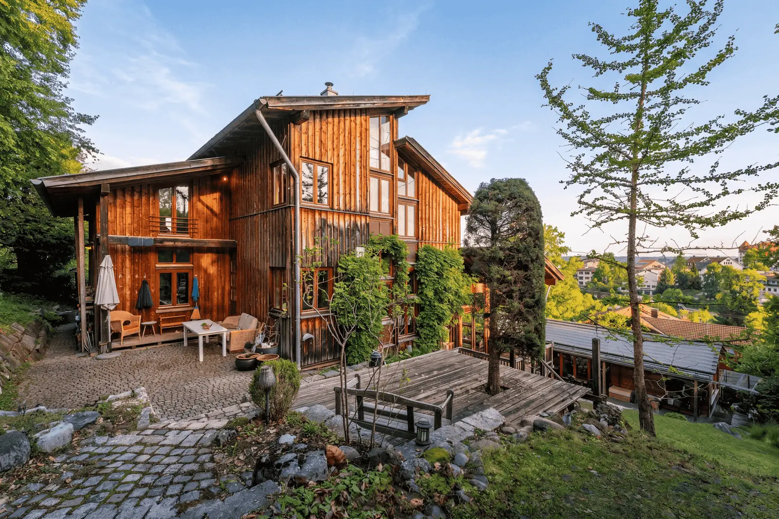 A large timber house with several terraces and outdoor seating, surrounded by trees and greenery on a sunny day.