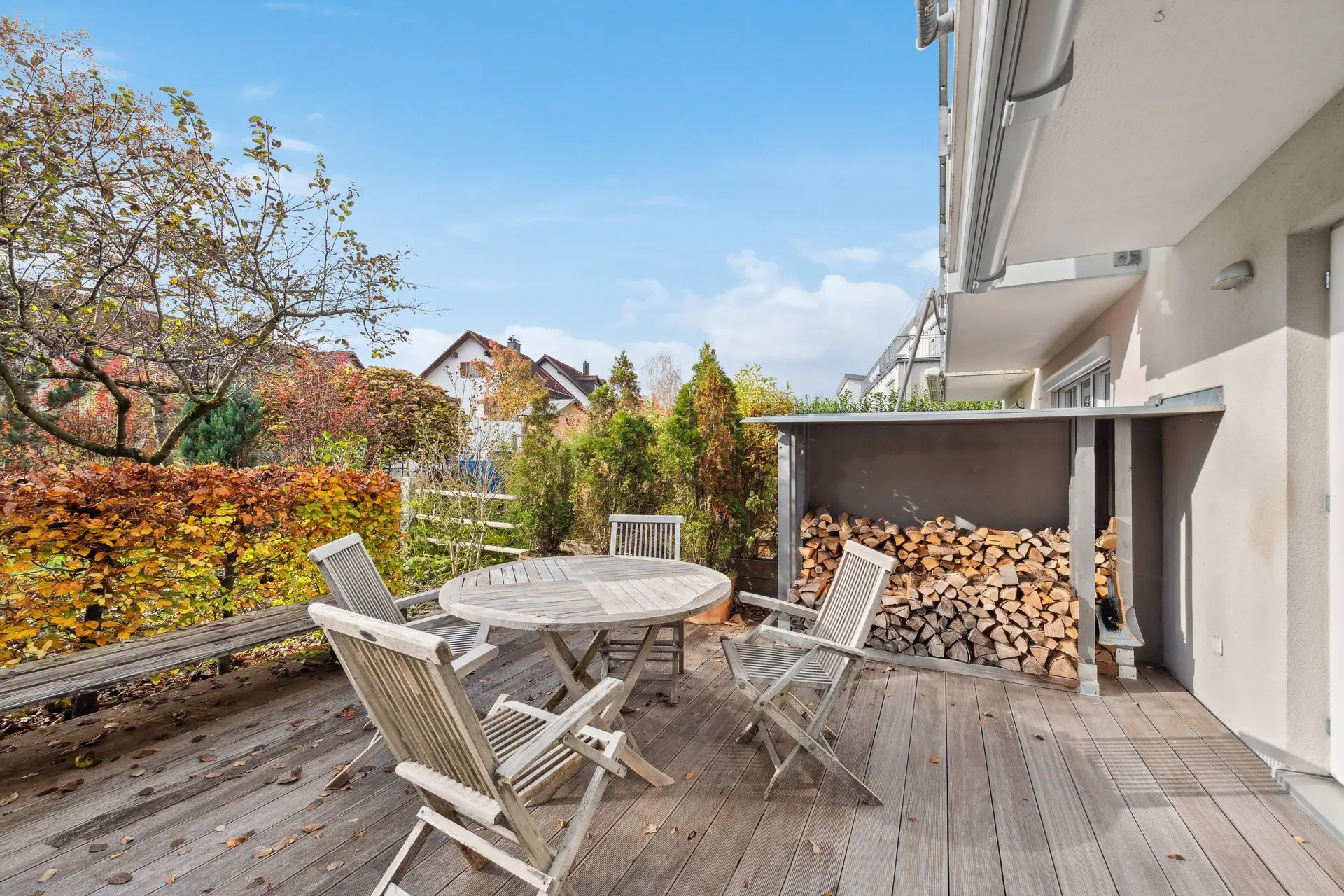 A wooden deck with a round table and four chairs, surrounded by autumn trees. A stack of firewood is neatly piled to the side.