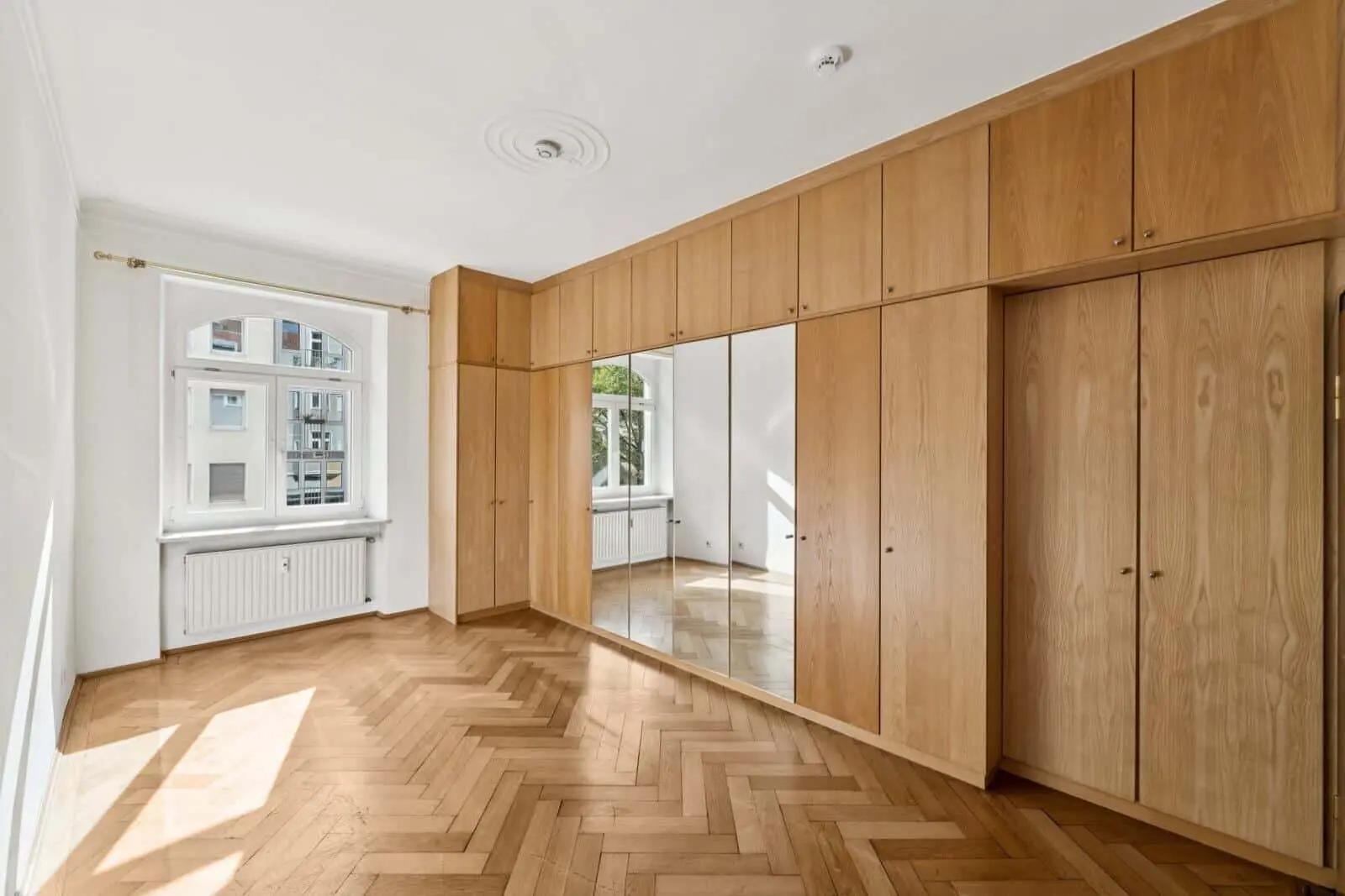 Bright empty room with herringbone wood floor, built-in wooden wardrobes with mirrors, large windows, and white walls.
